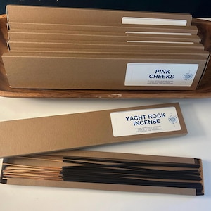 May include: A wooden bowl holds several brown cardboard boxes labeled "Pink Cheeks." Below, a box of "Yacht Rock Incense" is open, revealing incense sticks. Some incense sticks are black, others are brown.