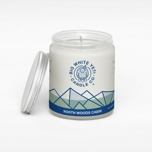 Pu&ograve; includere: Un barattolo di candela bianco con un design di montagna blu e verde e il testo "Big White Yeti Candle Co." e "North Woods Cabin".