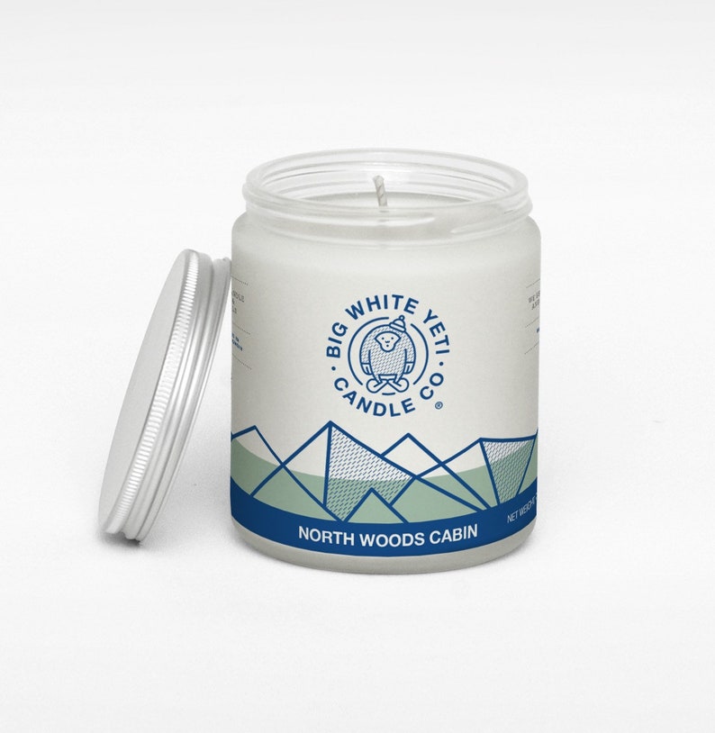 Pu&ograve; includere: Un barattolo di candela bianco con un design di montagna blu e verde e il testo "Big White Yeti Candle Co." e "North Woods Cabin".