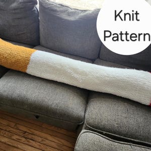 Puede incluir: Un gran cojín tejido en forma de cigarrillo en tonos amarillo, blanco, gris y rojo, apoyado en un sofá gris. El cojín tiene una punta amarilla, un cuerpo blanco y un extremo gris con una raya roja. El texto "Knit Pattern" está en un círculo blanco.