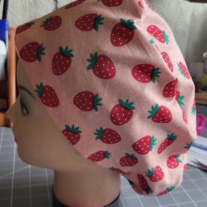 Peut inclure: Une charlotte chirurgicale avec un fond rose clair orné d'un motif répétitif de fraises rouges à feuilles vertes et de petits points blancs. Le bonnet présente une forme arrondie et froncée conçue pour couvrir la tête.