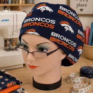 Peut inclure: Une charlotte chirurgicale bleu marine avec un motif répétitif du logo des Denver Broncos en blanc et orange, ainsi que le texte BRONCOS imprimé en lettres blanches et orange. La charlotte est présentée sur une tête de mannequin à lunettes.