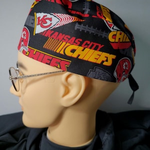 May include: Black scrub hat with a red, yellow, and white Kansas City Chiefs logo pattern. The hat has two ties.