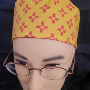 May include: A yellow scrub hat with a red and yellow pattern of the state of New Mexico's symbol. The hat is on a mannequin head wearing glasses.