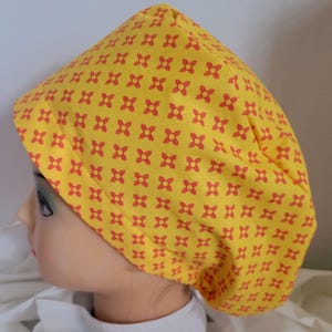 Peut inclure: Une charlotte chirurgicale jaune ornée d'un motif répétitif de petits symboles orange en forme de croix à quatre branches. Le bonnet présente une forme arrondie et ajustée avec une large bande repliée à la base, couvrant la tête.