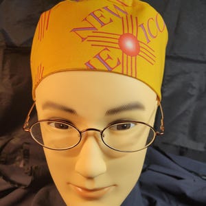 以下が含まれることがあります： ニューメキシコのデザインが施された黄色の手術用キャップ。州名と赤いジアシンボルが特徴です。 金色のフレームの楕円形の眼鏡をかけたマネキンが着用しています。