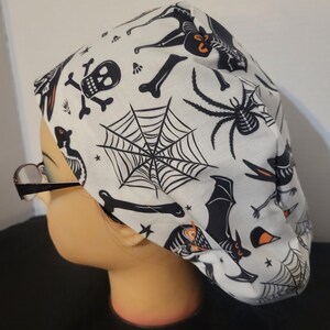 Peut inclure: Un bonnet de travail blanc avec un imprimé sur le thème d'Halloween représentant des chauves-souris, des araignées, des crânes, des os et des toiles d'araignées.