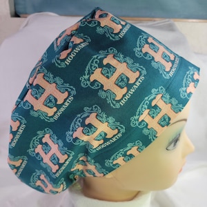 Può includere: Un cappello chirurgico blu-verde con un motivo ripetitivo dello stemma di Hogwarts e le parole "Hogwarts" in bianco e pesca.