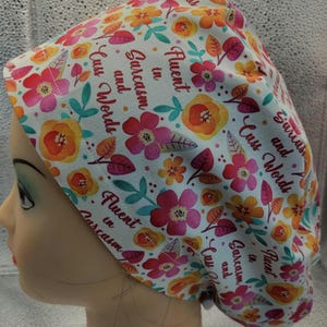 Peut inclure: Une charlotte de bloc opératoire à fond blanc ornée d'un motif floral coloré dans des tons rose, orange et bleu sarcelle. Le tissu est imprimé du texte répétitif "Fluent in Sarcasm and Cuss Words" en police cursive rouge foncé sur tout le motif.