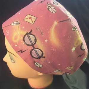 May include: A pink surgical scrub hat with a Harry Potter themed print. The print features glasses, a feather, a wand, a letter, a key, and stars.