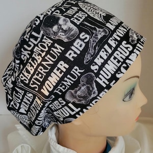 Peut inclure: Bonnet chirurgical noir et blanc avec un motif répété d'anatomie squelettique humaine. Le bonnet présente des images de crânes, de côtes et d'os, avec du texte tel que "CRÂNE", "CÔTES" et "SQUELETTE".