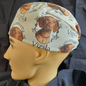 May include: A cream-colored surgical cap featuring a Vizsla dog portrait within a decorative frame. The cap is patterned with additional dog portraits and the text "Vizsla" and "Life's better with".