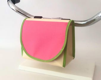 May include: A bicycle handlebar bag featuring a cream-coloured body with a vibrant pink flap and bright lime green trim. The bag is mounted on a silver metal handlebar and has a structured, rectangular shape with a rounded front flap closure.