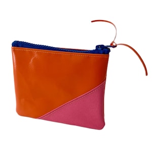Könnte beinhalten: Eine rechteckige Reißverschlusstasche mit einem glatten orangefarbenen oberen Teil und einem strukturierten rosa unteren Teil, die durch eine diagonale Naht getrennt sind. Die Tasche hat einen leuchtend blauen Reißverschluss mit einer dünnen orangefarbenen Lasche.