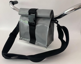 May include: A reflective gray roll-top bag with a black plastic buckle closure and a black adjustable shoulder strap, mounted on a chrome bicycle handlebar. The bag features a smooth, glossy finish and a vertical black nylon webbing strap.