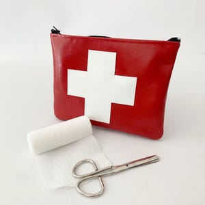 May include: A red first aid kit with a white cross, a roll of white gauze, and silver scissors. The kit has a black zip. The gauze is partially unrolled, and the scissors are open. The kit is a first aid essential.