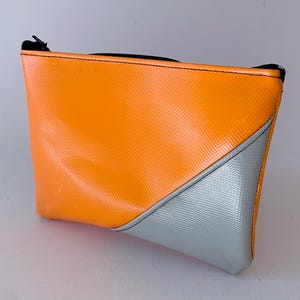 Puede incluir: Una pequeña bolsa con cremallera naranja y plateada. La bolsa está hecha de un material brillante y texturizado.