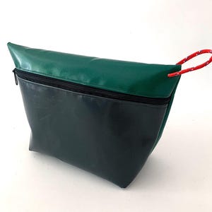Könnte beinhalten: Ein zweifarbiges Täschchen mit dunkelgrünem Oberteil und dunklerem grünem Boden. Das Täschchen hat einen schwarzen Reißverschluss und eine rot-weißes Seilschlaufe zum Tragen. Die Tasche ist aus strapazierfähigem, wasserdichtem Material gefertigt.