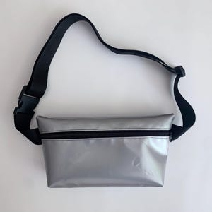 Könnte beinhalten: Eine silberne Bauchtasche mit einem schwarzen verstellbaren Riemen. Die Tasche hat einen Reißverschluss und eine glänzende Oberfläche.