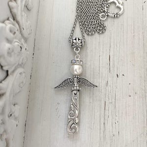 Op de afbeelding: Zilverkleurige ketting met een engelenvleugelhanger. De hanger heeft een engelontwerp met een parel en een decoratieve ketting. De ketting heeft een hartvormige sluiting. De achtergrond is een wit, rustiek houten oppervlak.