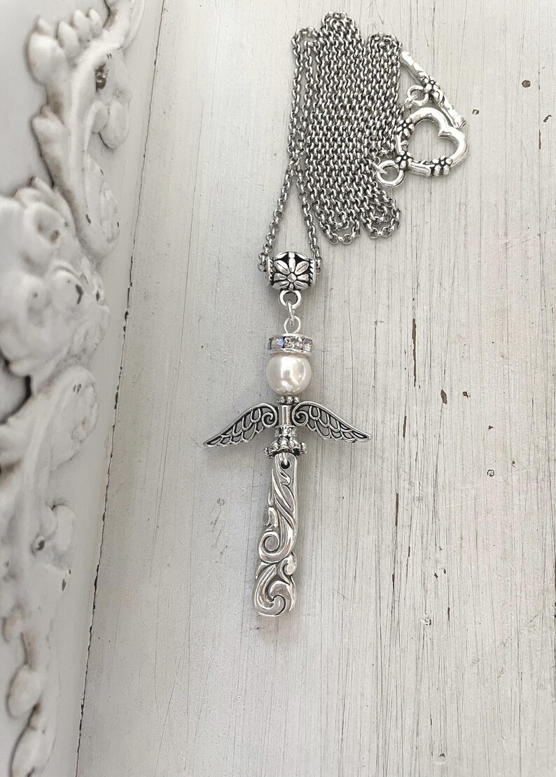 Op de afbeelding: Zilverkleurige ketting met een engelenvleugelhanger. De hanger heeft een engelontwerp met een parel en een decoratieve ketting. De ketting heeft een hartvormige sluiting. De achtergrond is een wit, rustiek houten oppervlak.
