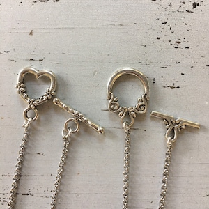 Peut inclure: Deux fermoirs &agrave; bascule de couleur argent&eacute;e pour la cr&eacute;ation de bijoux. Un fermoir pr&eacute;sente une boucle en forme de c&oelig;ur avec des d&eacute;tails floraux et une barre assortie. L'autre poss&egrave;de une boucle circulaire avec des volutes et une barre d&eacute;corative.