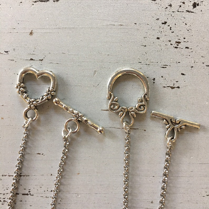 Peut inclure: Deux fermoirs &agrave; bascule de couleur argent&eacute;e pour la cr&eacute;ation de bijoux. Un fermoir pr&eacute;sente une boucle en forme de c&oelig;ur avec des d&eacute;tails floraux et une barre assortie. L'autre poss&egrave;de une boucle circulaire avec des volutes et une barre d&eacute;corative.