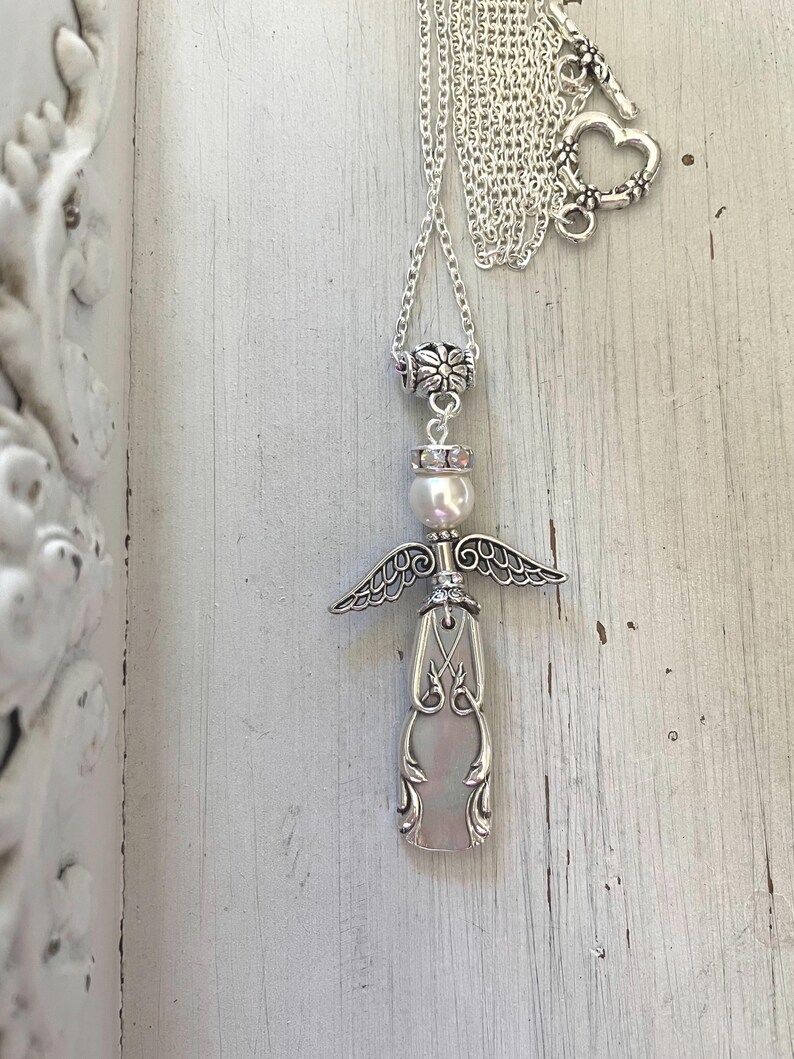 Peut inclure: Un collier argent&eacute; avec un pendentif en forme d'ange. Le pendentif est compos&eacute; d'un manche en m&eacute;tal d&eacute;coratif, d'une perle blanche en guise de t&ecirc;te et d'ailes en m&eacute;tal argent&eacute;. Le collier comprend une cha&icirc;ne argent&eacute;e avec un fermoir en c&oelig;ur.
