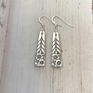 May include: A pair of silver-toned dangle earrings featuring a tapered, flat rectangular shape. The surface displays an embossed floral pattern at the base and a vertical, chevron-like leaf design extending upward to the hook-style ear wire.