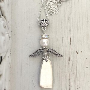 May include: A silver-toned angel pendant necklace. The necklace features a detailed angel charm with outstretched wings, a pearl, and a toggle clasp with a heart design. The chain is delicate and the pendant is approximately 2 inches in length.