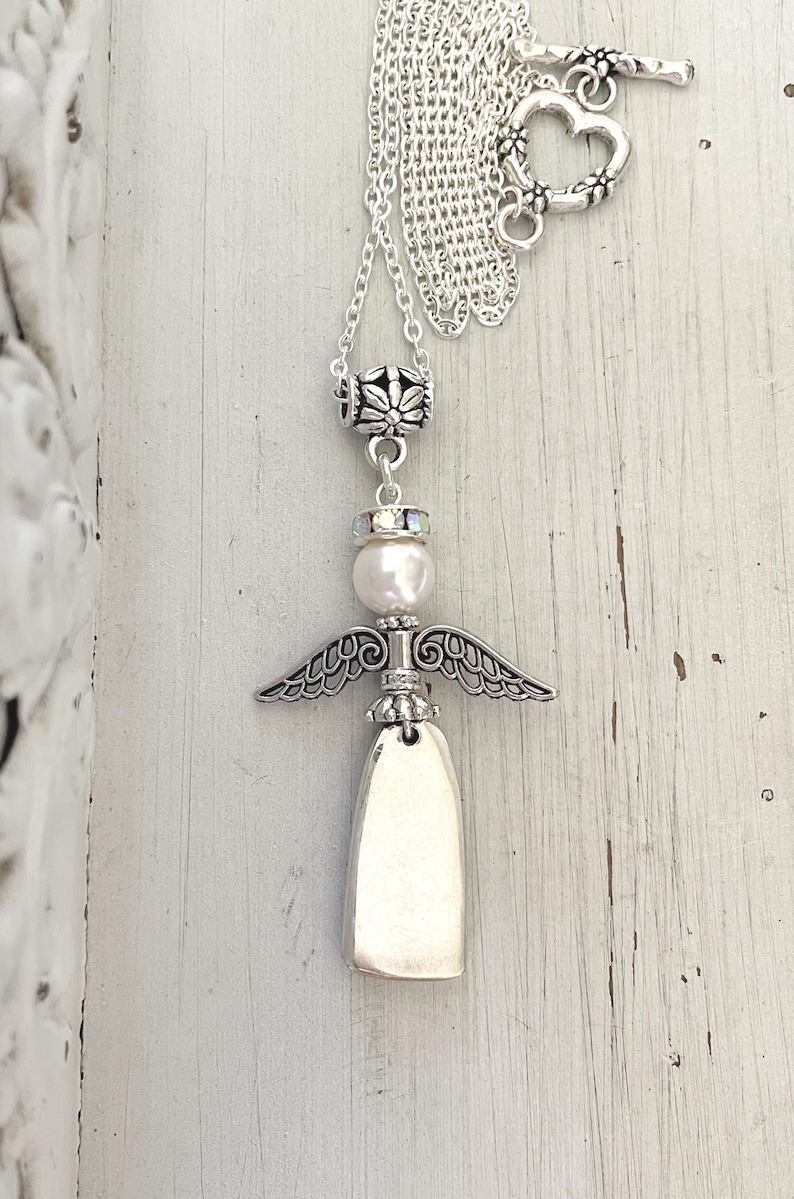 May include: A silver-toned angel pendant necklace. The necklace features a detailed angel charm with outstretched wings, a pearl, and a toggle clasp with a heart design. The chain is delicate and the pendant is approximately 2 inches in length.