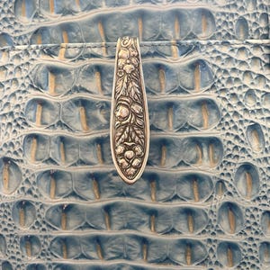 May include: Close-up of a light blue leather handbag with a silver clasp. The clasp is intricately designed with floral and leaf patterns. The bag's surface has a textured, crocodile-like pattern.