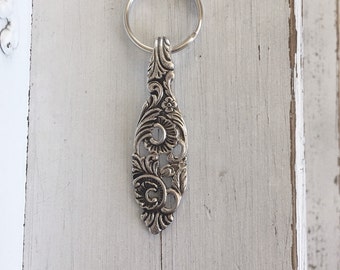 May include: Silver-colored, ornate spoon-shaped keychain. The keychain features a decorative floral and scroll design. The spoon-shaped pendant is attached to a silver metal key ring. The background is a white, distressed wooden surface.