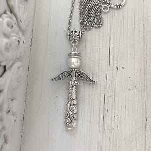 Op de afbeelding: Zilverkleurige engel hanger ketting. De hanger heeft een engel ontwerp met een parel hoofd, gedetailleerde vleugels en een sierlijk, krullend lichaam. De ketting heeft een delicate ketting met een hartvormige sluiting.