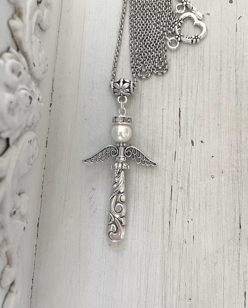 Op de afbeelding: Zilverkleurige engel hanger ketting. De hanger heeft een engel ontwerp met een parel hoofd, gedetailleerde vleugels en een sierlijk, krullend lichaam. De ketting heeft een delicate ketting met een hartvormige sluiting.