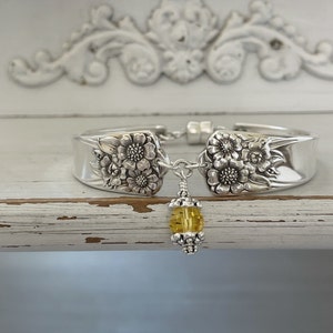May include: A silver bracelet crafted from repurposed silverware. The bracelet features floral designs on the spoon heads and a dangling yellow bead. The bracelet is connected with a chain and magnetic clasp.