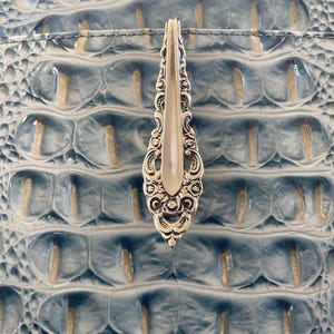 May include: A silver-toned metal spoon handle featuring an ornate, embossed floral and scrollwork pattern at the base, positioned vertically against a textured, light blue surface with a repeating oval pattern resembling reptile skin.