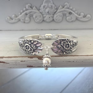 May include: A silver-toned bracelet crafted from two curved spoon handles featuring an embossed floral pattern. The two ends are connected by a magnetic clasp, and a single white pearl bead charm hangs from a silver-toned metal setting in the center.