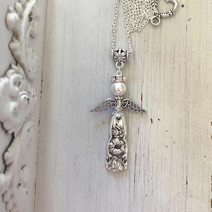 Peut inclure: Un collier argent&eacute; avec un pendentif en forme d'ange. Le pendentif se compose d'une perle blanche ronde pour la t&ecirc;te, d'ailes en m&eacute;tal et d'une base en m&eacute;tal textur&eacute; &agrave; motif floral. Le collier comprend une fine cha&icirc;ne argent&eacute;e.