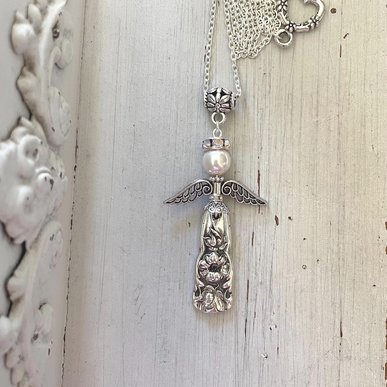 Peut inclure: Un collier argent&eacute; avec un pendentif en forme d'ange. Le pendentif se compose d'une perle blanche ronde pour la t&ecirc;te, d'ailes en m&eacute;tal et d'une base en m&eacute;tal textur&eacute; &agrave; motif floral. Le collier comprend une fine cha&icirc;ne argent&eacute;e.