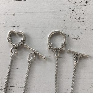 May include: Close-up of three silver-toned toggle clasps. One is heart-shaped with floral detailing, another is a circular design with ornate swirls, and the third has a decorative bar. Each clasp has a chain attached.