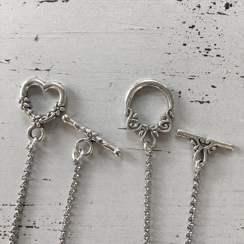 May include: Close-up of three silver-toned toggle clasps. One is heart-shaped with floral detailing, another is a circular design with ornate swirls, and the third has a decorative bar. Each clasp has a chain attached.