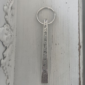 May include: A silver-colored keychain with a rectangular pendant. The pendant is adorned with an engraved floral pattern. The keychain ring is circular. The background is a light gray, distressed wooden surface.