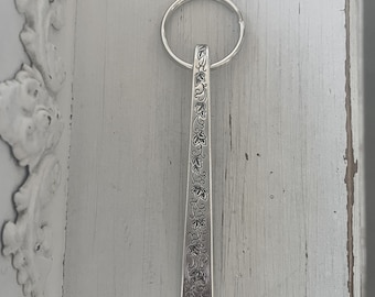 May include: A silver-colored keychain with a rectangular pendant. The pendant is adorned with an engraved floral pattern. The keychain ring is circular. The background is a light gray, distressed wooden surface.
