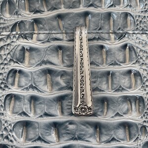 May include: A silver-toned metal spoon handle featuring an intricate embossed floral pattern along the center and a larger flower motif at the base. The handle has a smooth, polished finish with decorative scalloped edges running down its length.