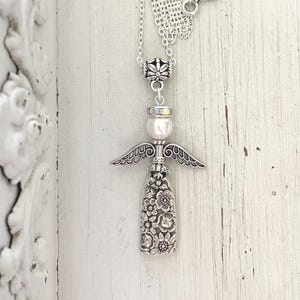 May include: Silver-toned angel pendant necklace. The pendant features an angel design with detailed wings, a pearl, and a floral-patterned body. The necklace has a toggle clasp with a heart-shaped design.