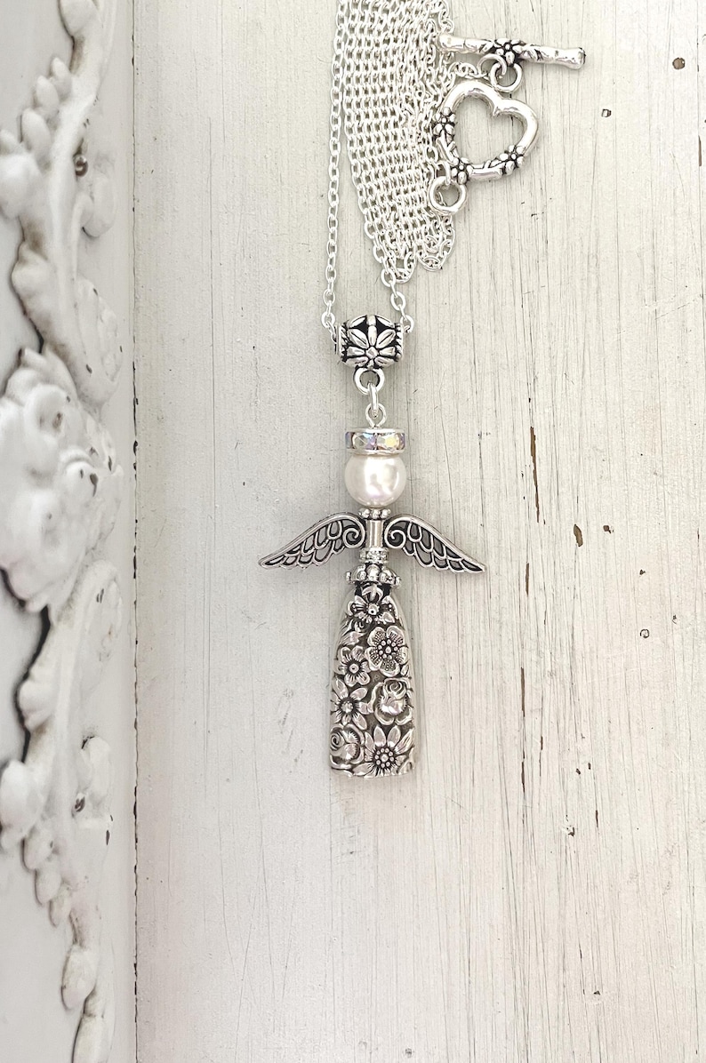 May include: Silver-toned angel pendant necklace. The pendant features an angel design with detailed wings, a pearl, and a floral-patterned body. The necklace has a toggle clasp with a heart-shaped design.