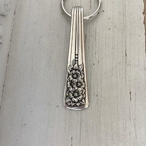 May include: Silver-toned metal keychain featuring a floral design. The keychain is made from a repurposed spoon, with a decorative floral pattern on the handle. The key ring is attached to the top of the spoon handle.