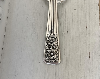 May include: Silver-toned metal keychain featuring a floral design. The keychain is made from a repurposed spoon, with a decorative floral pattern on the handle. The key ring is attached to the top of the spoon handle.