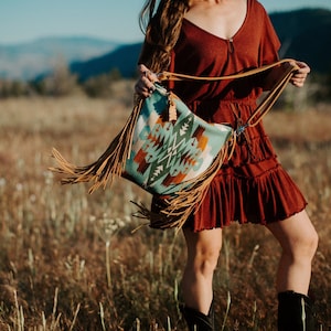May include: A turquoise and brown patterned shoulder bag with long brown fringe. The bag has a leather strap and a decorative tassel. The bag is being held in a field of tall grass.